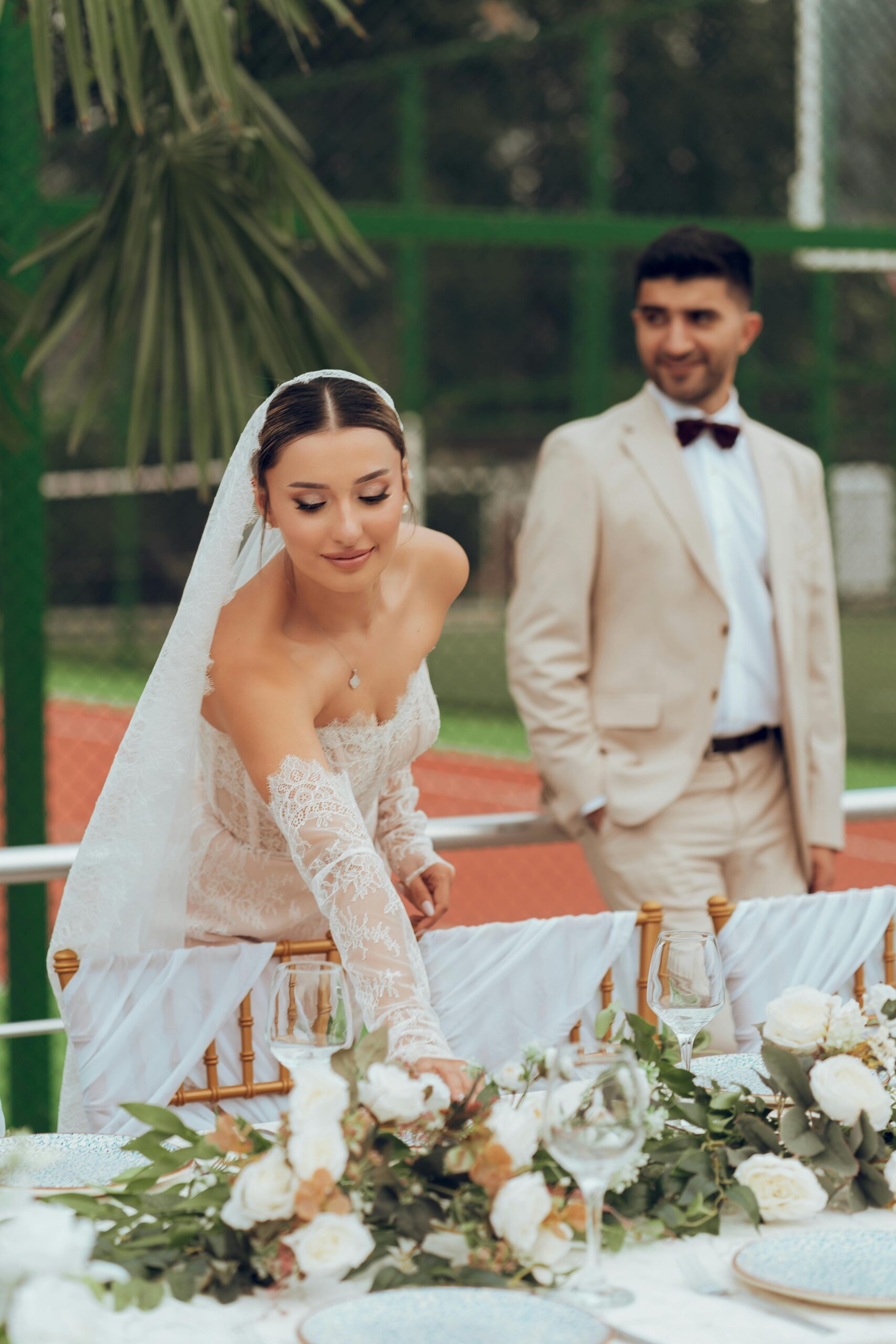 A bride, dressed in an elegant lace wedding gown and veil, gracefully arranges a flower bouquet on a table